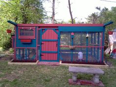 a red and blue chicken coop sitting on top of a lush green field next to a park bench