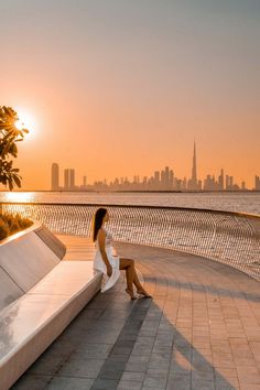 a woman sitting on a bench in front of the ocean at sunset with cityscape in the background