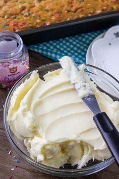 a bowl filled with white frosting next to a pan of cake and sprinkles