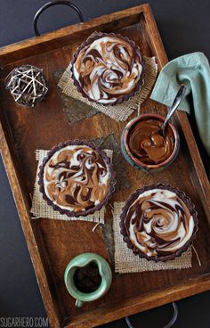 three pies with chocolate and marshmallow toppings on a wooden tray next to a cup of coffee