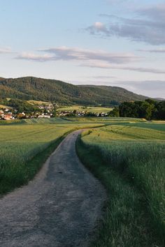 an empty dirt road in the middle of a green field with houses and hills in the distance