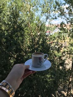 a person holding a coffee cup and saucer in their left hand, with trees in the background