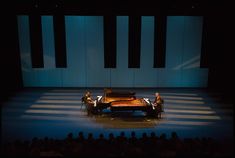 two men sitting at a piano on stage in front of an audience