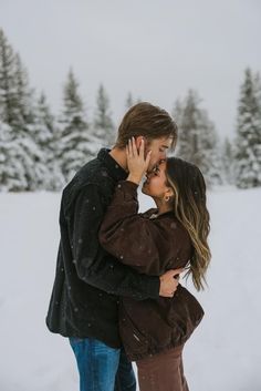 a man and woman kissing in the snow with pine trees behind them on a snowy day