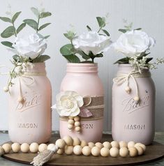 three pink mason jars with white flowers in them on a wooden tray next to beads