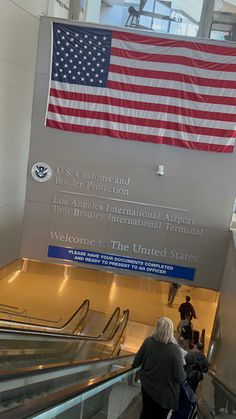 an american flag hanging from the ceiling above escalators in a building with people walking up and down them