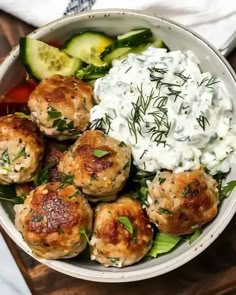 a white bowl filled with meatballs, cucumbers and sour cream on top of a wooden table