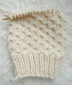 a white knitted hat laying on top of a pile of fluffy white fur next to a pair of knitting needles