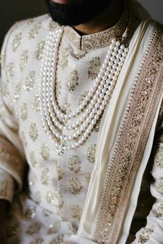 a man in a white and gold wedding outfit with pearls on his neck, standing