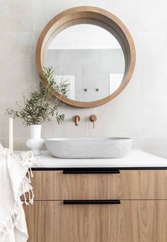 a white sink sitting under a round mirror on top of a wooden cabinet next to a plant