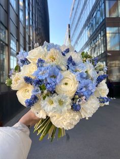 a bouquet of white and blue flowers is held by a woman's hand in front of tall buildings