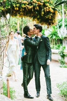 two men in suits kissing each other under an arch with flowers hanging from the ceiling