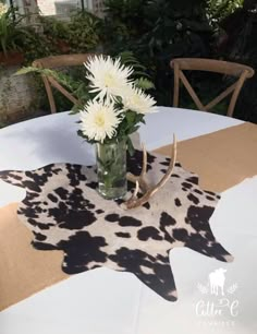 a vase filled with white flowers sitting on top of a cow print table cloth next to a deer's head