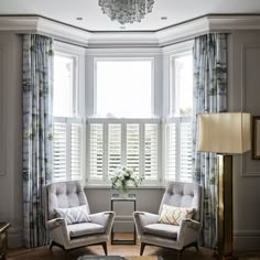 two chairs sitting in front of a window with shutters on each side and a chandelier hanging from the ceiling