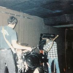 two young men are playing instruments in front of a speaker and guitar player, while another man stands behind them