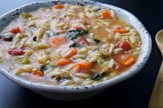 a white bowl filled with pasta and vegetables next to a wooden spoon on a table