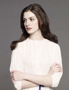 a woman with her arms crossed standing in front of a gray background wearing a white blouse