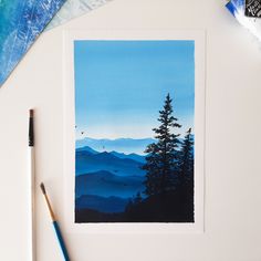 a watercolor painting of mountains and trees on a white table with paintbrushes