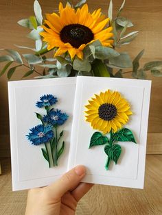 two cards with sunflowers and blue flowers on them are held in front of a vase