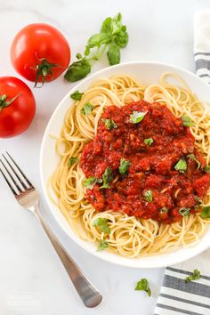 a white bowl filled with spaghetti and tomato sauce next to two forks, tomatoes and basil on the side