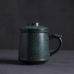 a dark green ceramic teapot on a gray surface with the lid open and handle extended