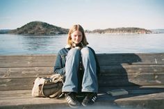 a woman sitting on a wooden bench next to the water with a book bag in front of her