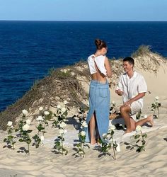 a man and woman sitting on top of a sandy beach next to the ocean with flowers