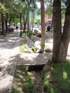 a small stream running through a park next to trees and people walking on the sidewalk