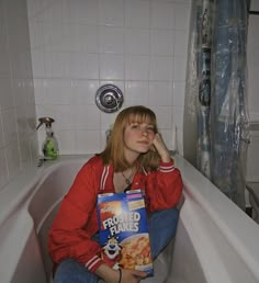 a woman sitting on the edge of a bathtub holding a box of cereal