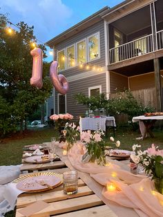 an outdoor table set up for a party with candles and flowers on it, in front of a house