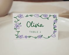 a place card sitting on top of a white plate with purple and green flowers around it