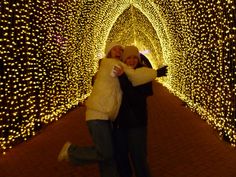 two people standing in front of a tunnel covered with christmas lights at the end of a walkway