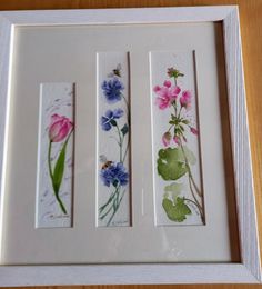 three framed pictures with flowers painted on them in white frames, sitting on a wooden table