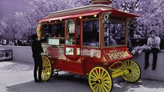 a man standing next to a red and yellow cart in front of a tree filled with purple flowers