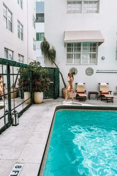 a woman standing next to a swimming pool in front of a white building with green shutters