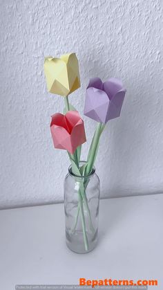 three origami flowers in a vase on a white table with a wall behind them