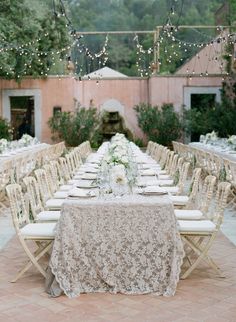 a long table set up with chairs and tables covered in white linens, surrounded by string lights