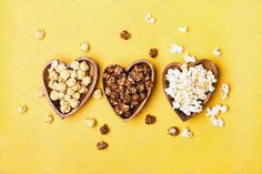 three wooden heart shaped bowls filled with popcorn and nuts on a yellow background, top view