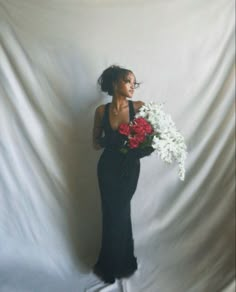 a woman in a long black dress holding white and red flowers with her hands behind her back