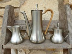 three silver coffee pots sitting on top of a wooden shelf next to a stone wall