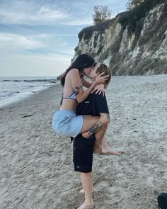 two people are hugging on the beach with one person standing up and another sitting down