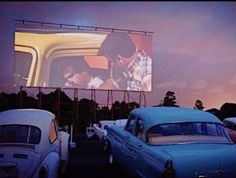 an old car is parked in front of a large screen with two people on it