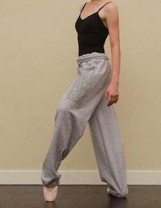 a woman standing in front of a wall with her legs crossed and wearing grey sweatpants