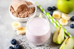 a smoothie with blueberries and bananas next to it on a gray surface, surrounded by other fruits