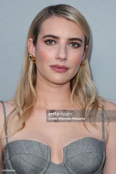 a woman with long blonde hair wearing a gray dress and gold hoop earrings, standing in front of a grey wall