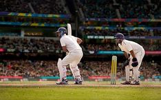 two men in white uniforms playing a game of cricket on a field with people watching from the stands