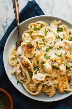 a white bowl filled with pasta and chicken on top of a blue cloth next to a fork