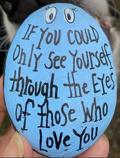 a person holding up a painted rock with words on it and a cat in the background