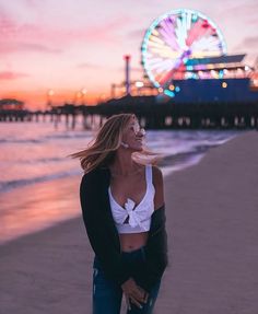 a woman standing on top of a sandy beach next to the ocean at sunset with a ferris wheel in the background