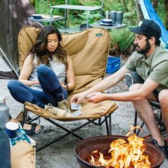 a man and woman sitting in chairs next to a campfire